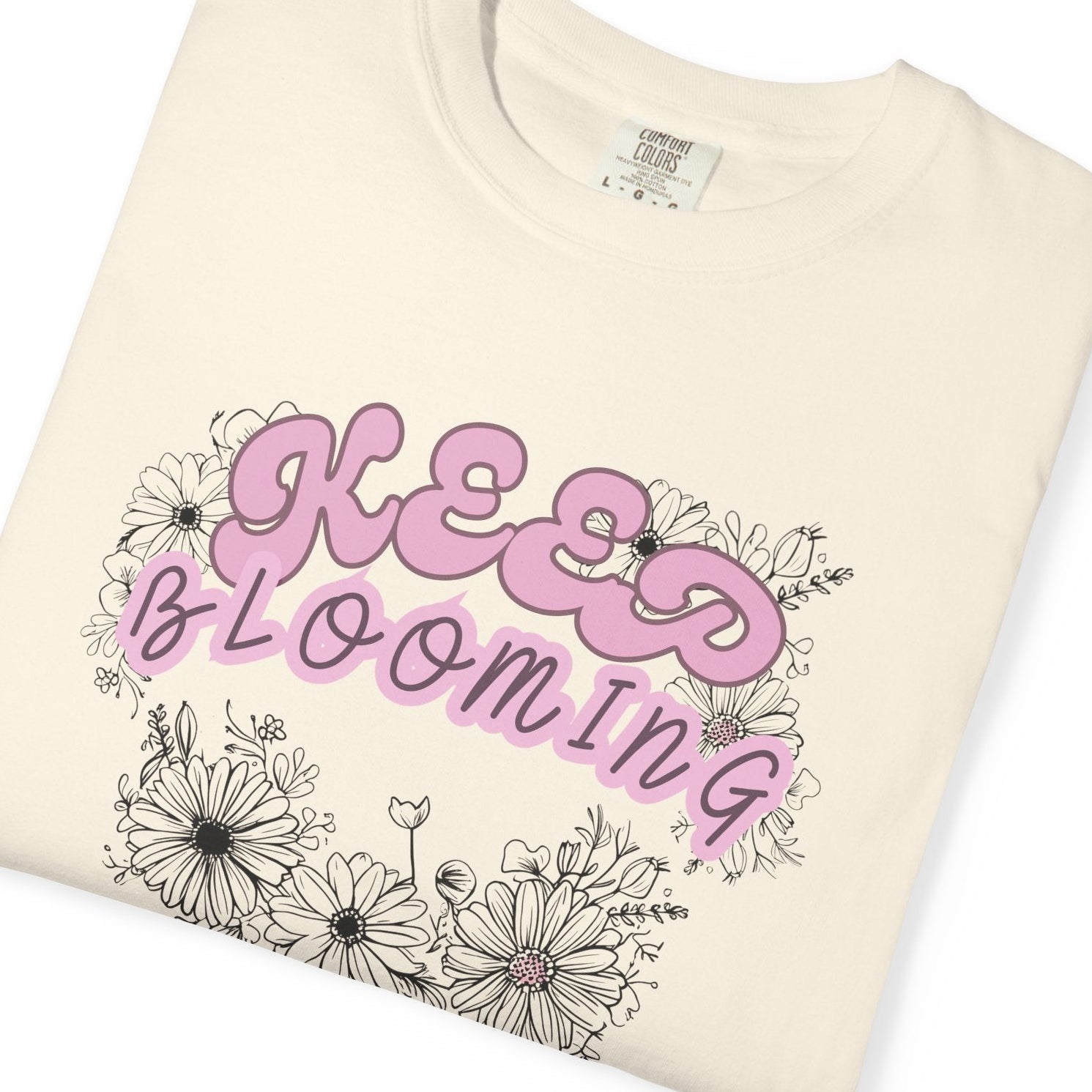 A folded beige t-shirt with a floral design and the text 'KEEP BLOOMING' in pink above the floral pattern.
